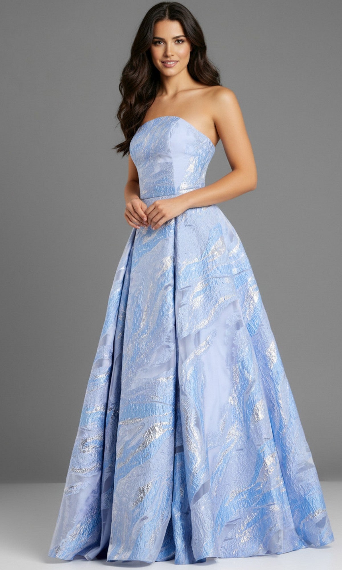 A woman wears the Jovani 44369 long prom dress in blue, featuring a strapless brocade design with silver patterns, standing against a plain gray background.